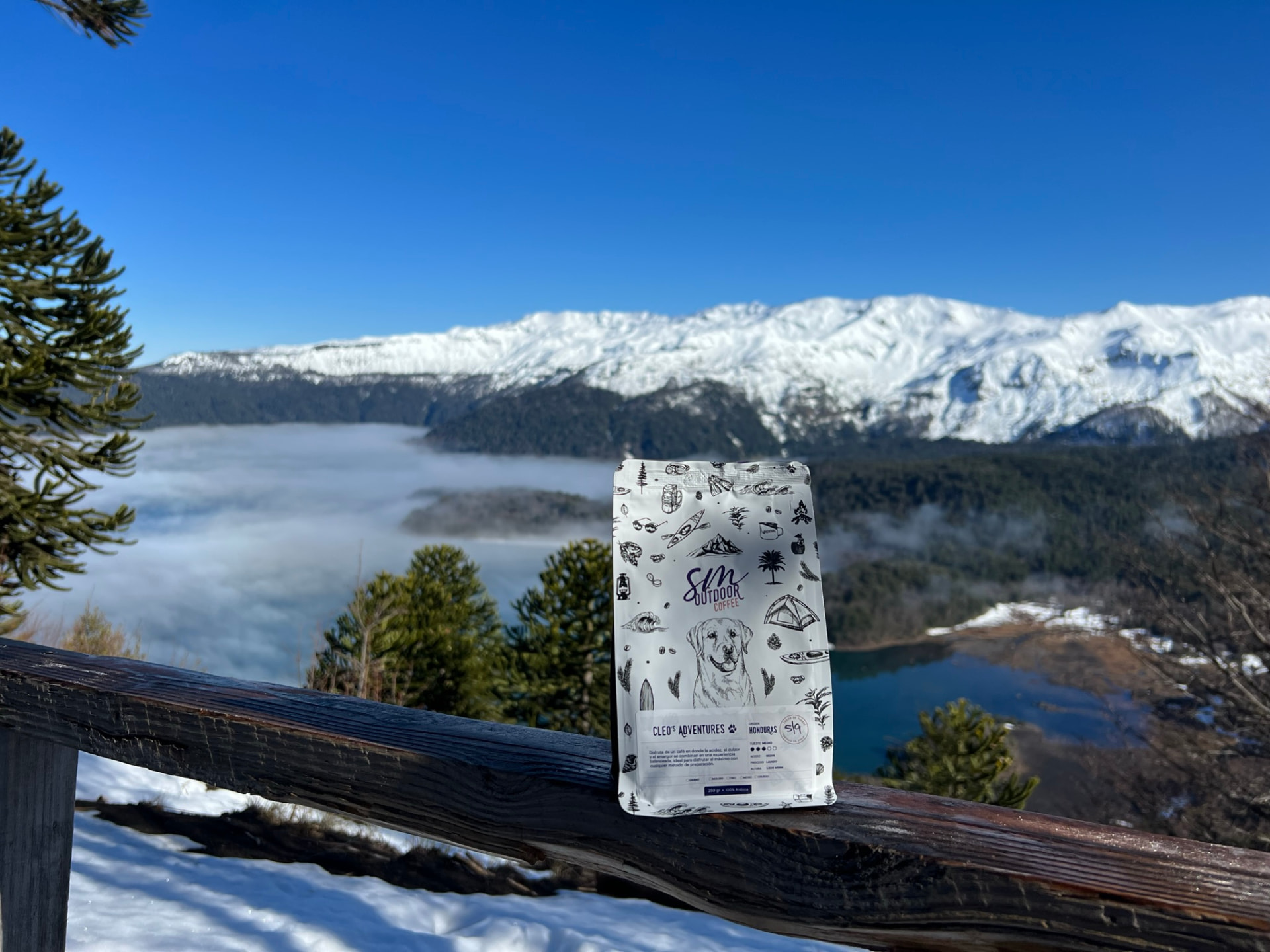Bolsa SM Outdoor Coffee Cleo's Adventures sobre una baranda de madera frente a montañas nevadas y valle con niebla