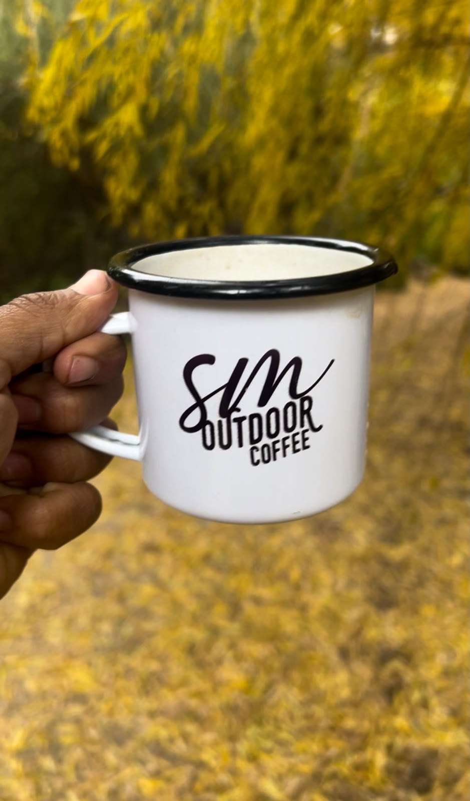 Taza enlozada blanca con el logo SM Outdoor Coffee sostenida en la mano al aire libre